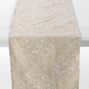 A beige table runner adorned with an intricate white floral and geometric pattern is draped over a white table.