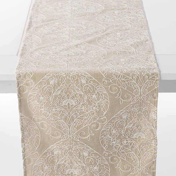 A beige table runner adorned with an intricate white floral and geometric pattern is draped over a white table.