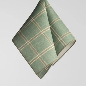 A green and beige plaid fabric napkin folded and hanging against a plain light grey background.