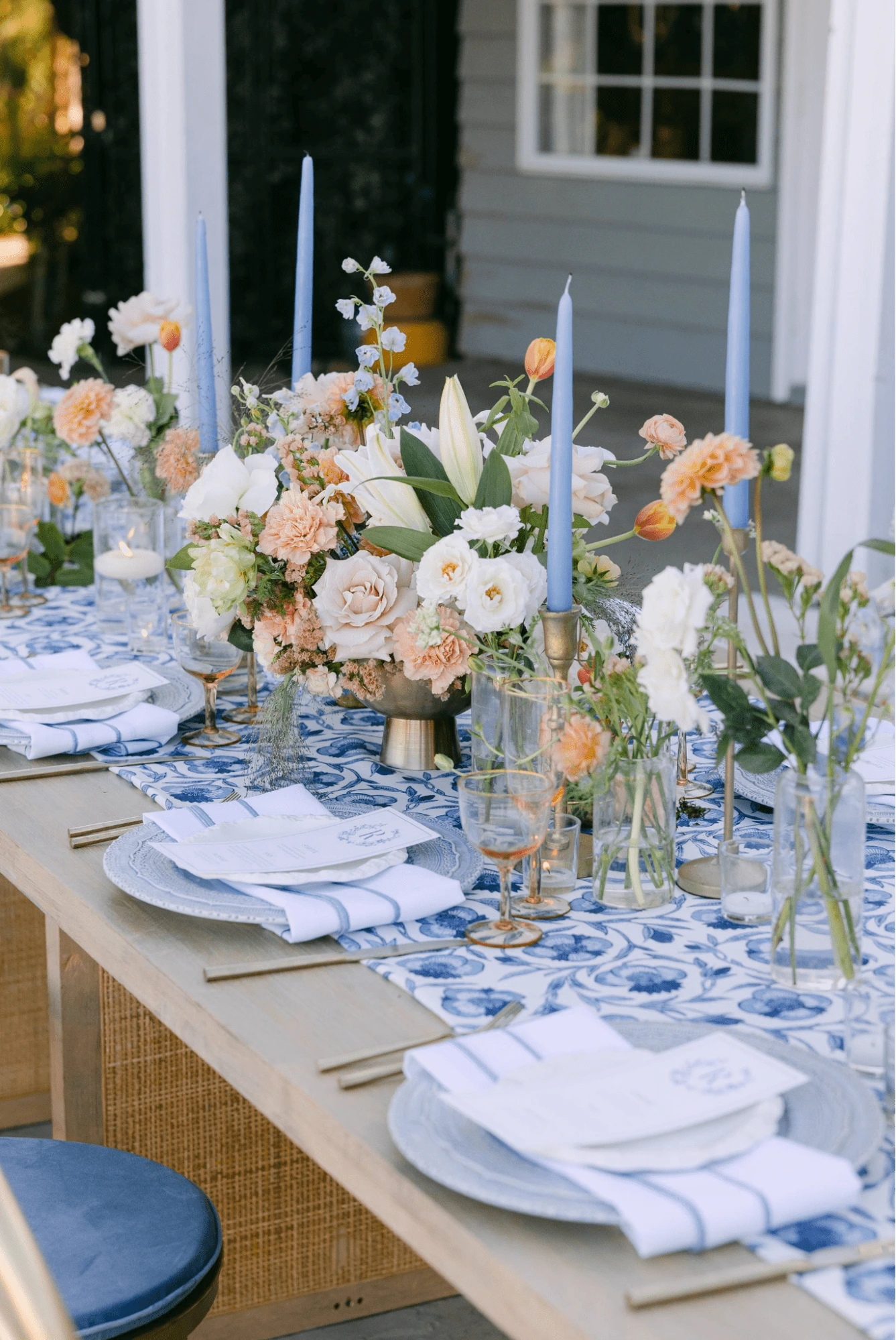 A decorated dining table with blue candles, floral arrangements, and blue-patterned napkins set outside by a house.