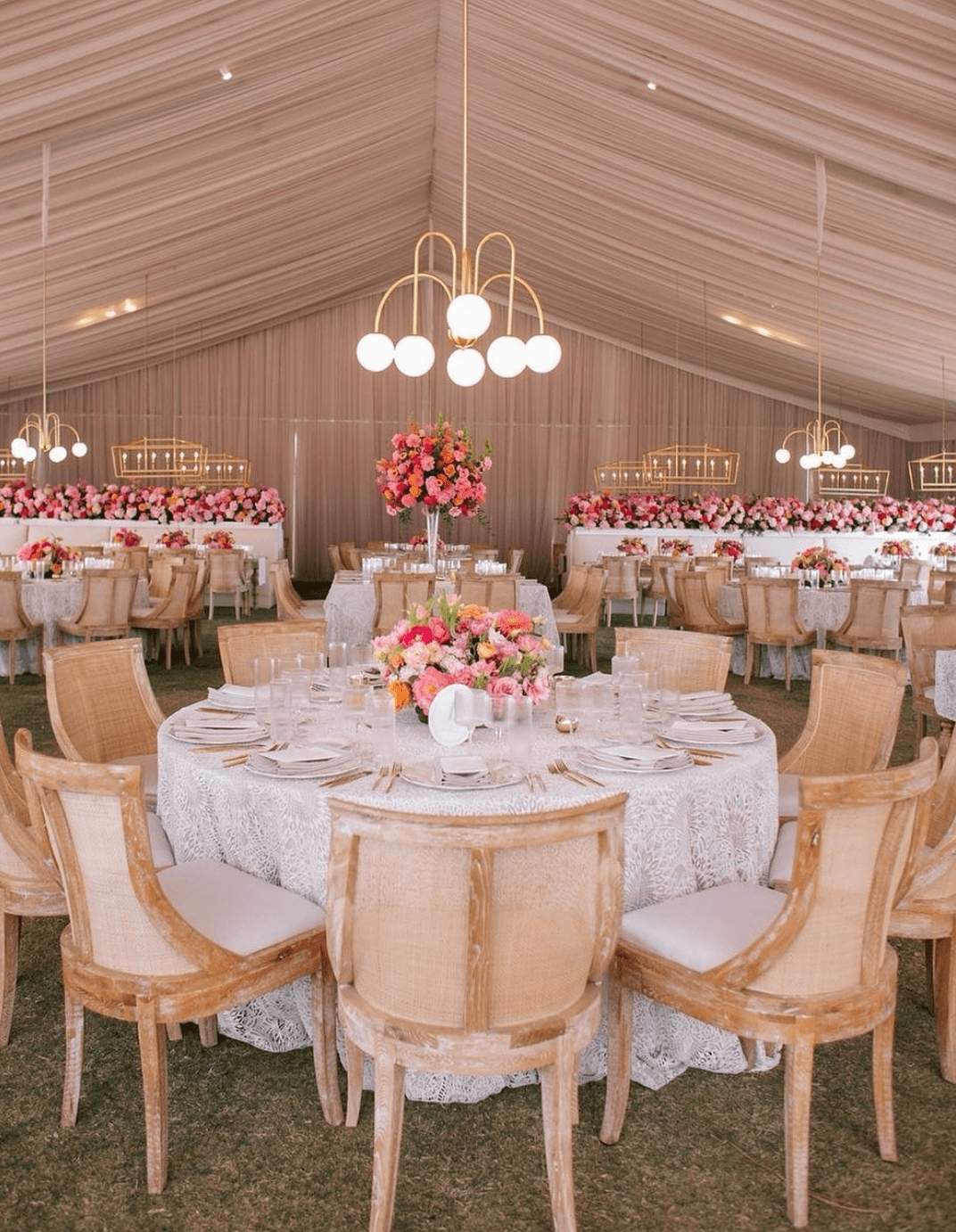 Large tented reception area with elegant round tables set with white lace tablecloths, pink floral centerpieces, and beige wooden chairs. Hanging lights and additional floral arrangements in the background.