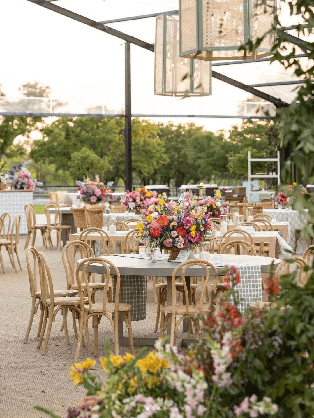 Outdoor event space with wooden chairs and tables set with floral centerpieces. Overhead lighting and greenery surround the area.