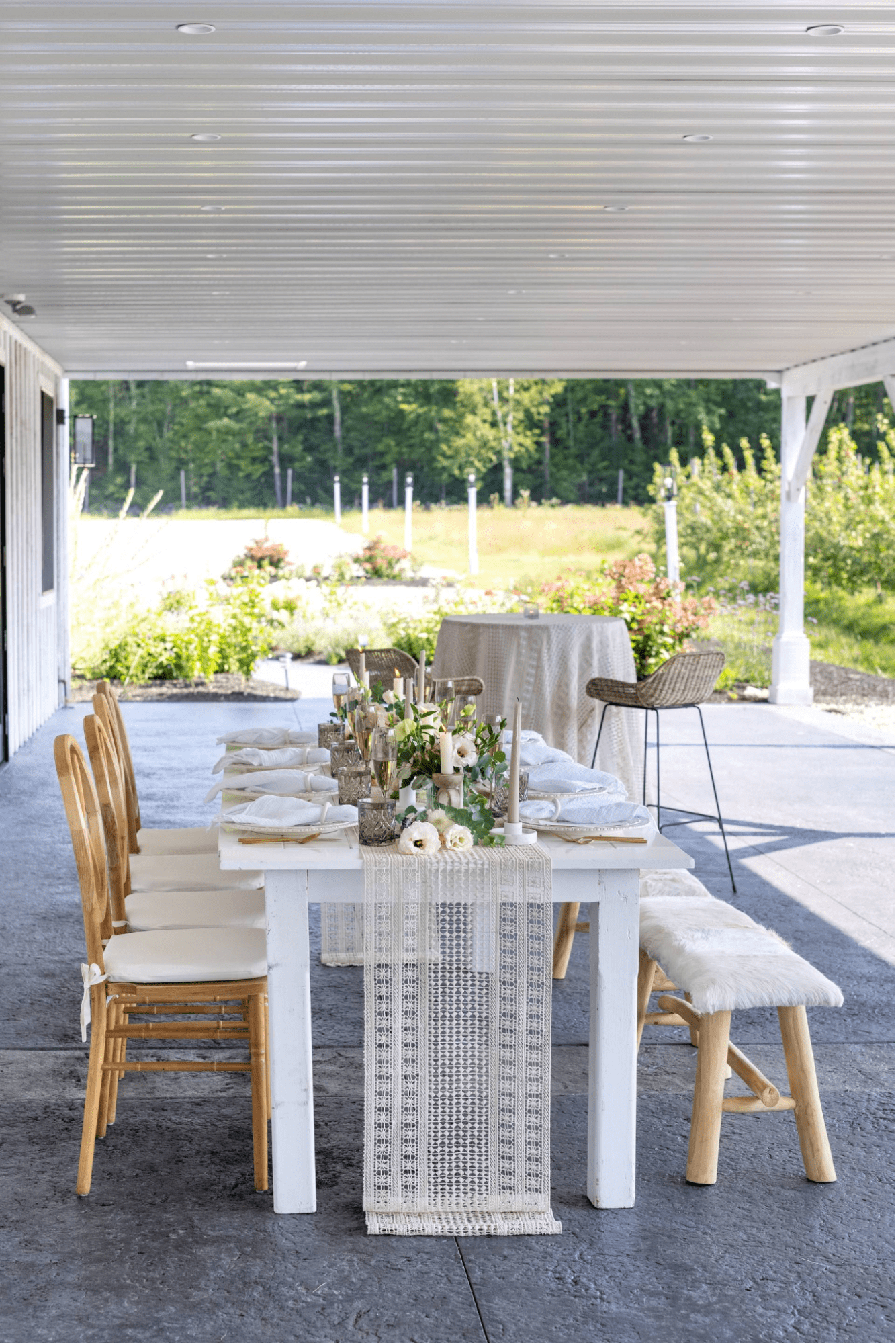 A long white table with a lace runner is set for a meal under a covered patio. Wooden chairs with white cushions surround the table. In the background, a round table and bar stool are visible.