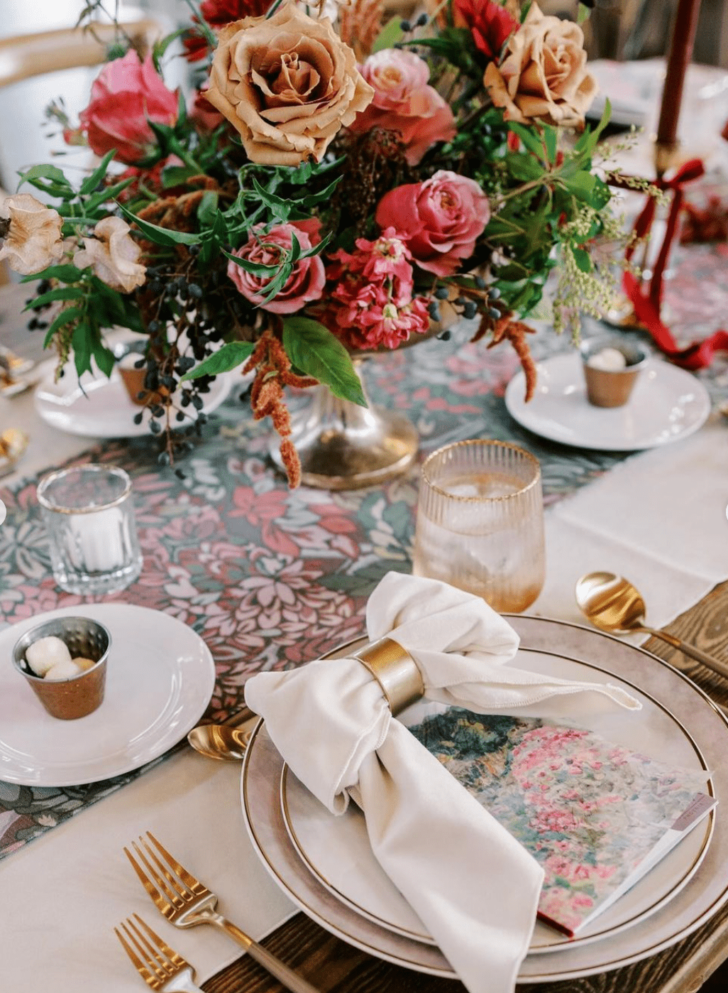 A table set with elegant dinnerware, gold utensils, and a floral arrangement centerpiece. A white napkin with a gold napkin ring is placed on a plate featuring a colorful floral print.