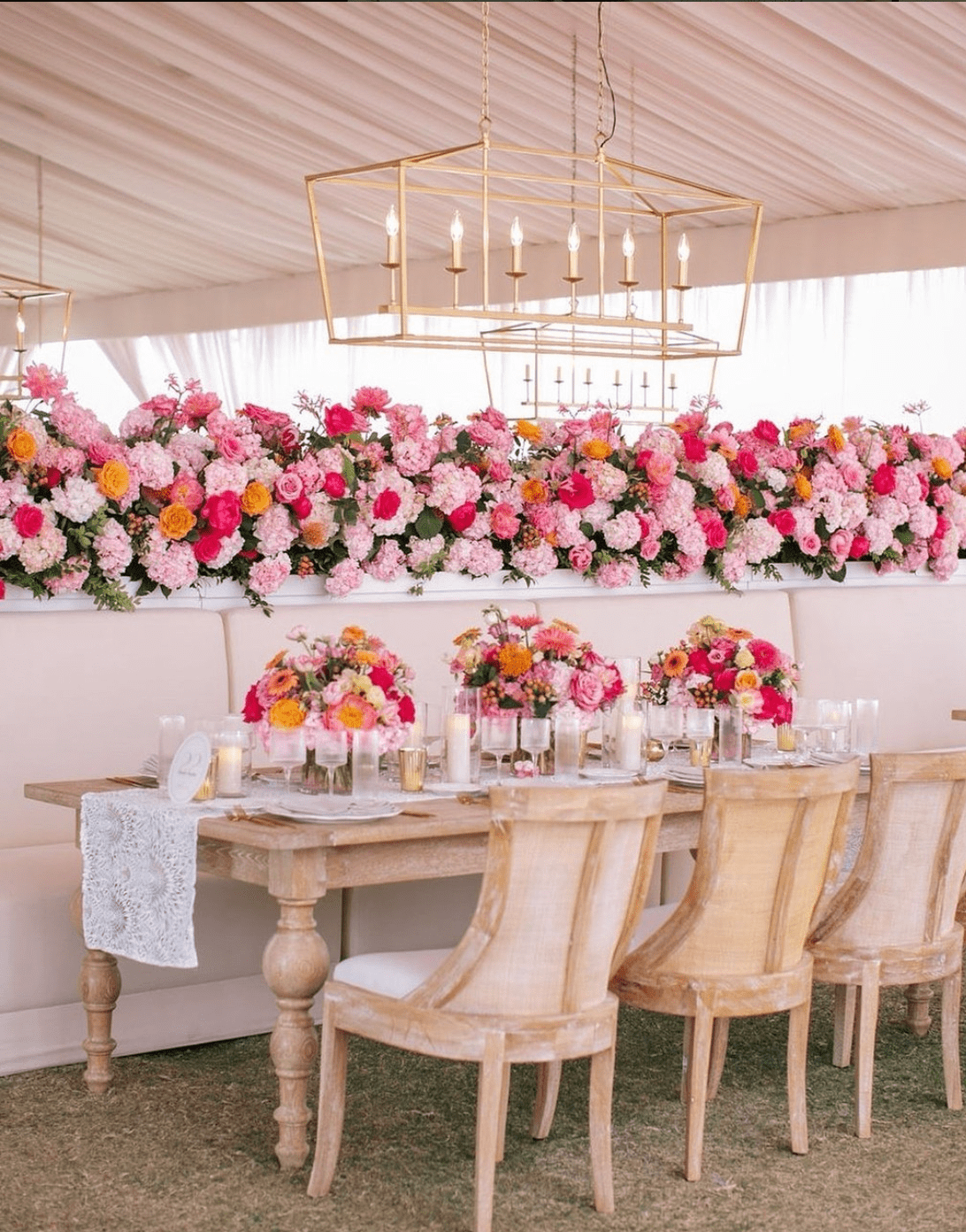 A wooden table and chairs are set up in an event tent, decorated with pink, orange, and white floral arrangements and lit by contemporary gold chandeliers.