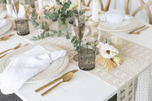 A white table is set with gold cutlery, textured glassware, white plates with cloth napkins, and decorated with flowers, greenery, and candles.