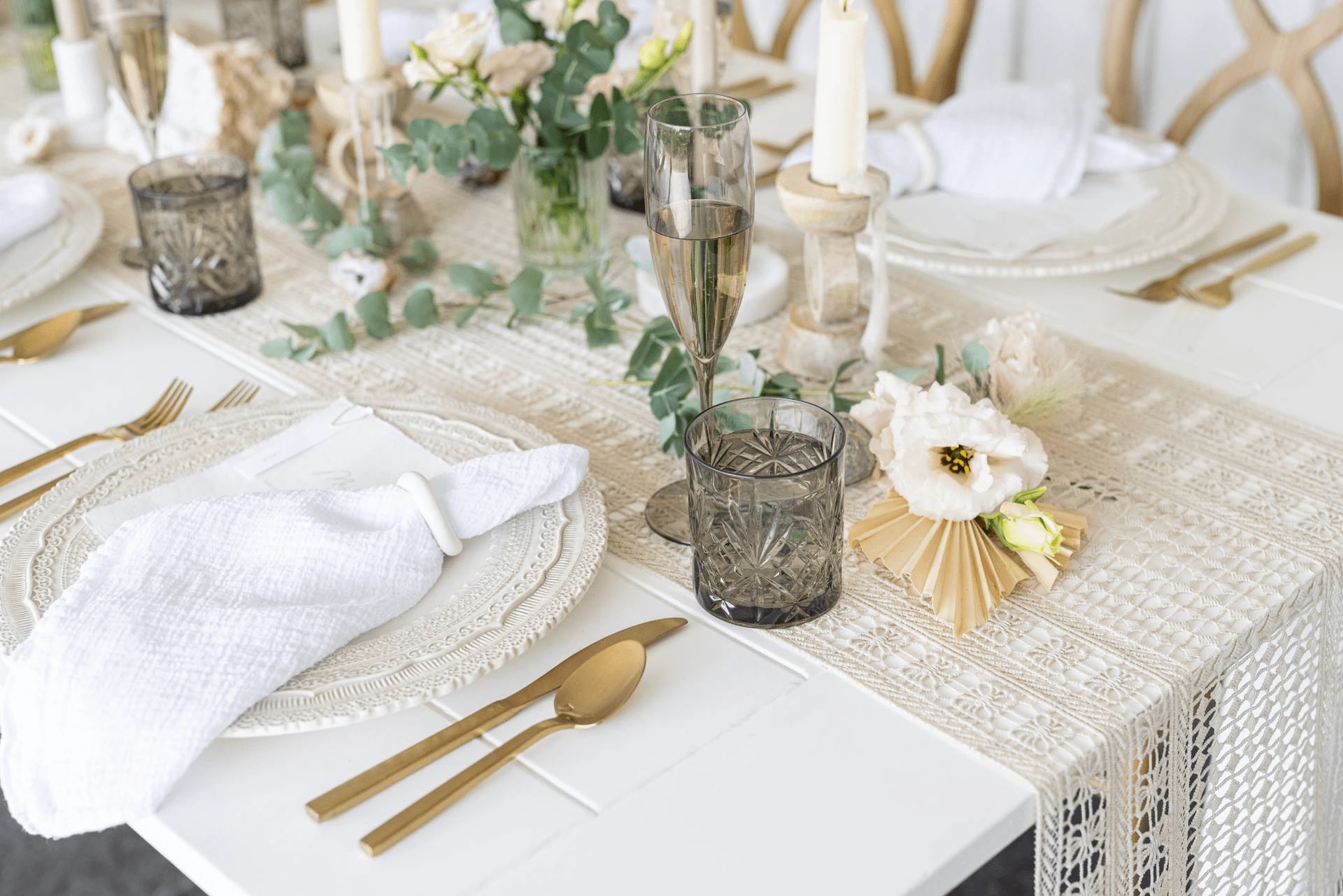 A white table is set with gold cutlery, textured glassware, white plates with cloth napkins, and decorated with flowers, greenery, and candles.