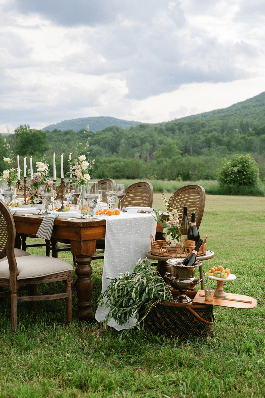 An outdoor dining setup on a grassy field with a table set for a meal, including flowers, candles, and tableware, surrounded by chairs. In the background, there is a scenic view of green hills and cloudy sky.