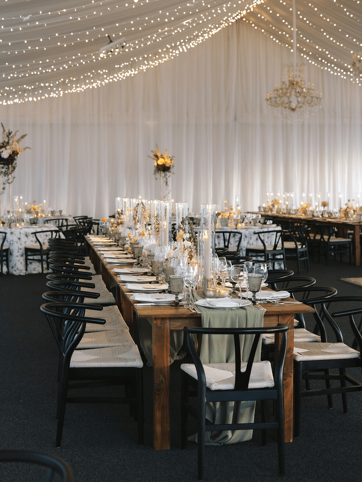 Long wooden tables set for dinner under a tent with string lights and chandeliers, featuring black chairs and elegant place settings with tall candles and floral centerpieces.