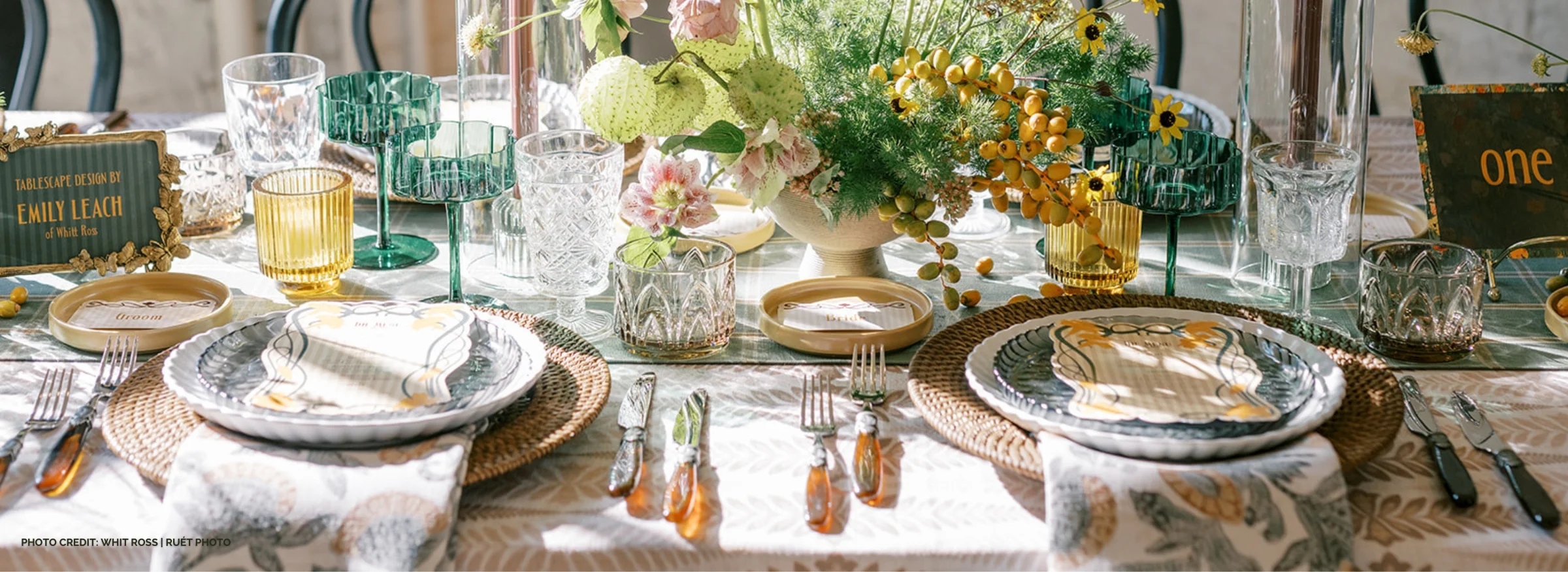 A table is set for a meal with ornate dishes, glasses, and flatware. Centerpieces of flowers and greenery adorn the table, which also showcases place cards, decorative napkins, and elegant linens provided by our table linen rental service.