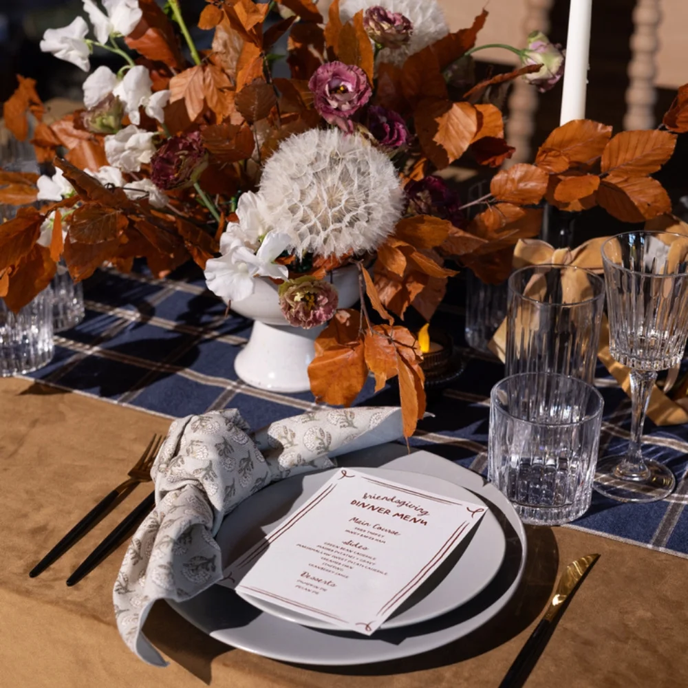 A table setting with a floral centerpiece and a menu card on a plate. Crystal glasses and utensils are arranged on a blue and brown tablecloth.