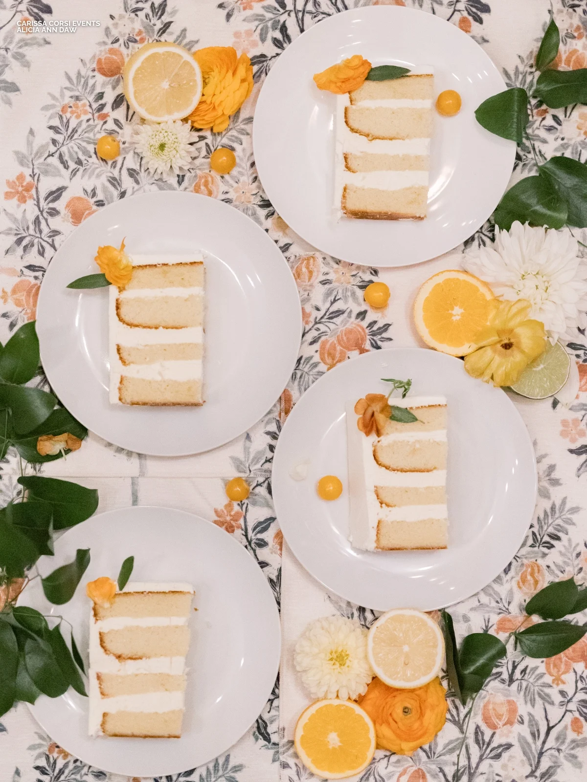 Four plates of layered cake slices with cream filling, garnished with orange flowers and decorated with citrus fruit and green leaves on a floral tablecloth.