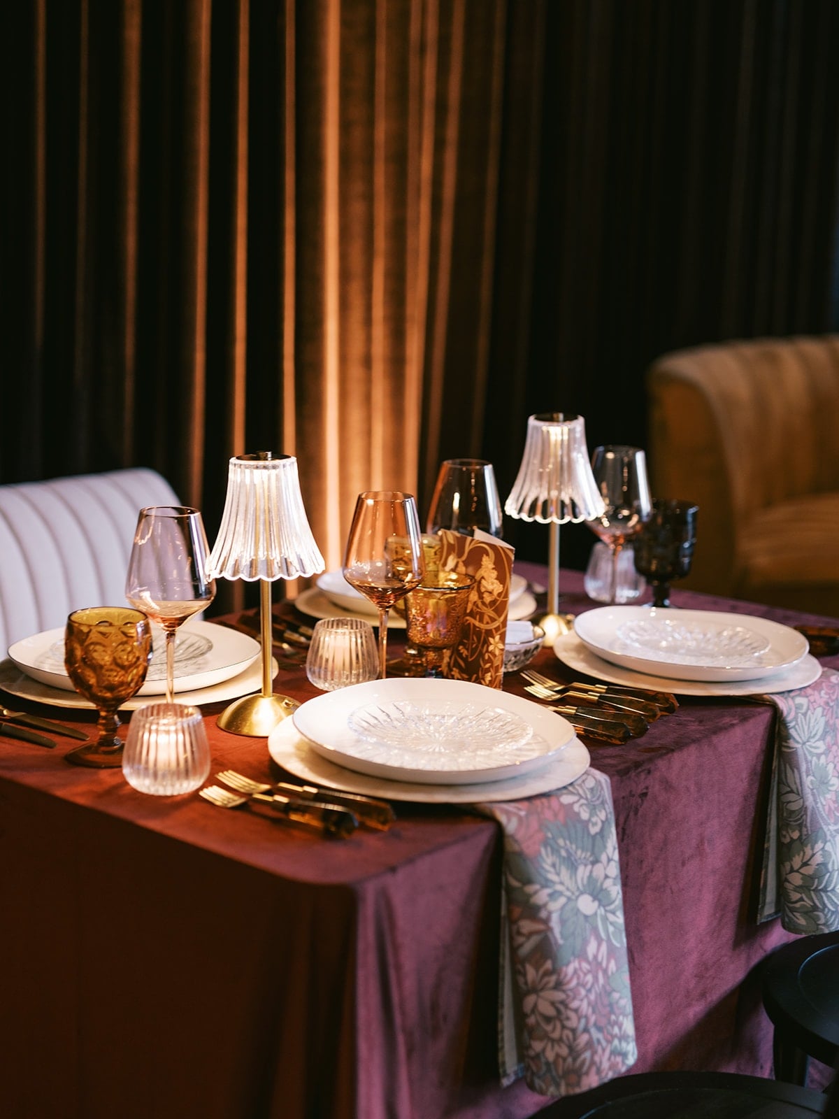 Elegant dining table set with white plates, assorted glasses, silverware, and decorative lamps on a burgundy tablecloth, featuring floral patterned edges.