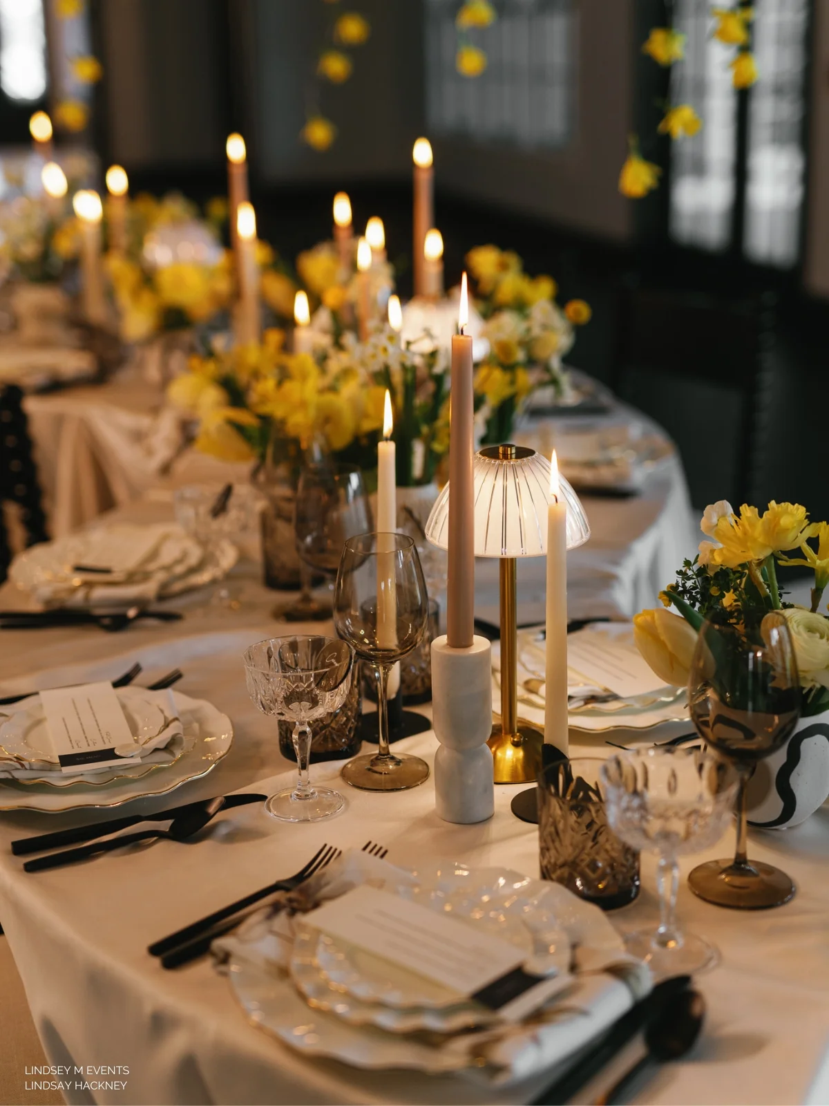 A beautifully set dinner table with lit candles, elegant tableware, and yellow floral arrangements.