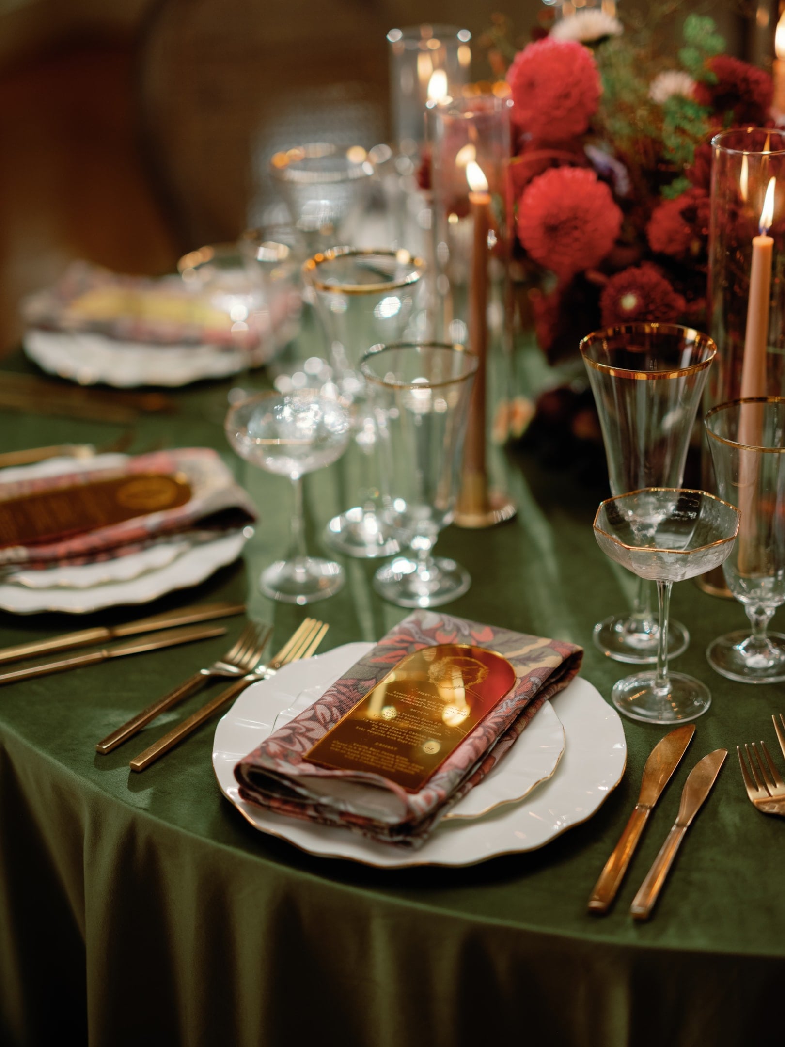 A formal dining table with green tablecloth, gold cutlery, glassware, candles, and a floral centerpiece. Plates are set with decorative napkins.