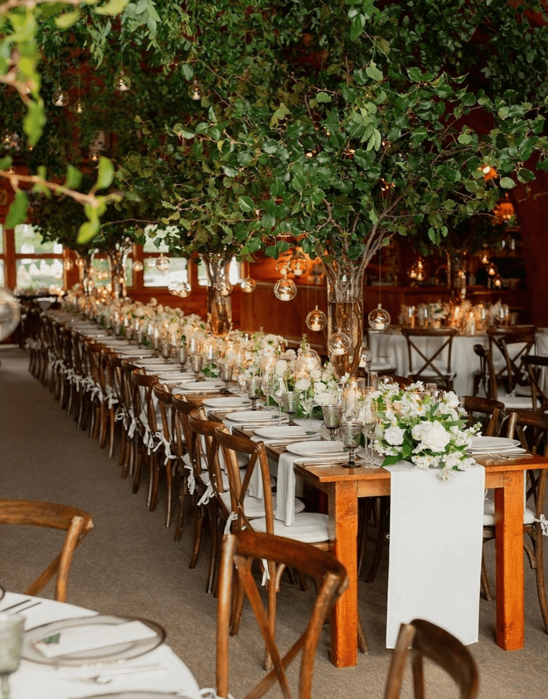 Long dining table set for an event, decorated with white flowers, lush greenery, and hanging bulbs, surrounded by wooden chairs in a rustic venue.