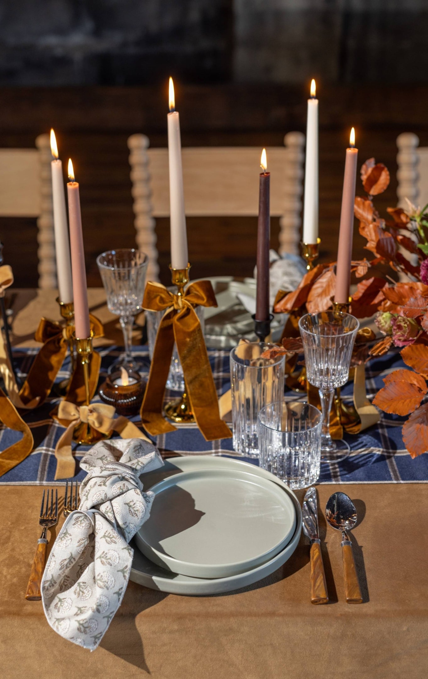 A dining table set with plates, cutlery, glasses, and tall lit candles in holders. Napkins are elegantly folded, and autumn-themed floral arrangements are present.