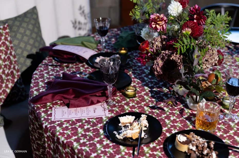 A table set for a meal with floral-patterned tablecloth, wine glasses, plates of desserts, and a bouquet of flowers in the center.