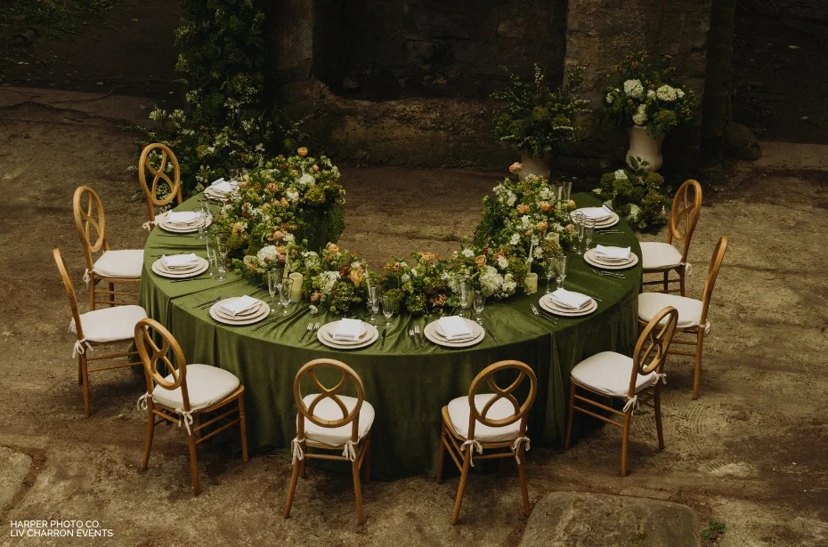 A semi-circular table set for eight with green tablecloth, white plates, and floral centerpieces. Seven wooden chairs surround the table in an outdoor setting.