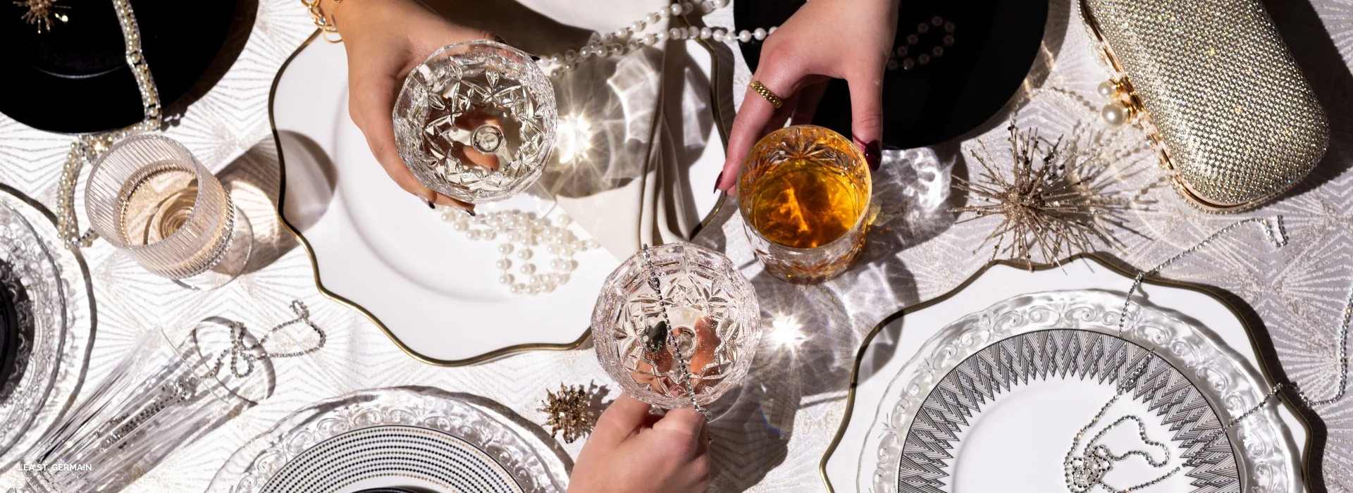 Two hands holding crystal glasses over an elegant table set with ornate plates, decorative items, and pearls, accentuated by exquisite linen rentals.