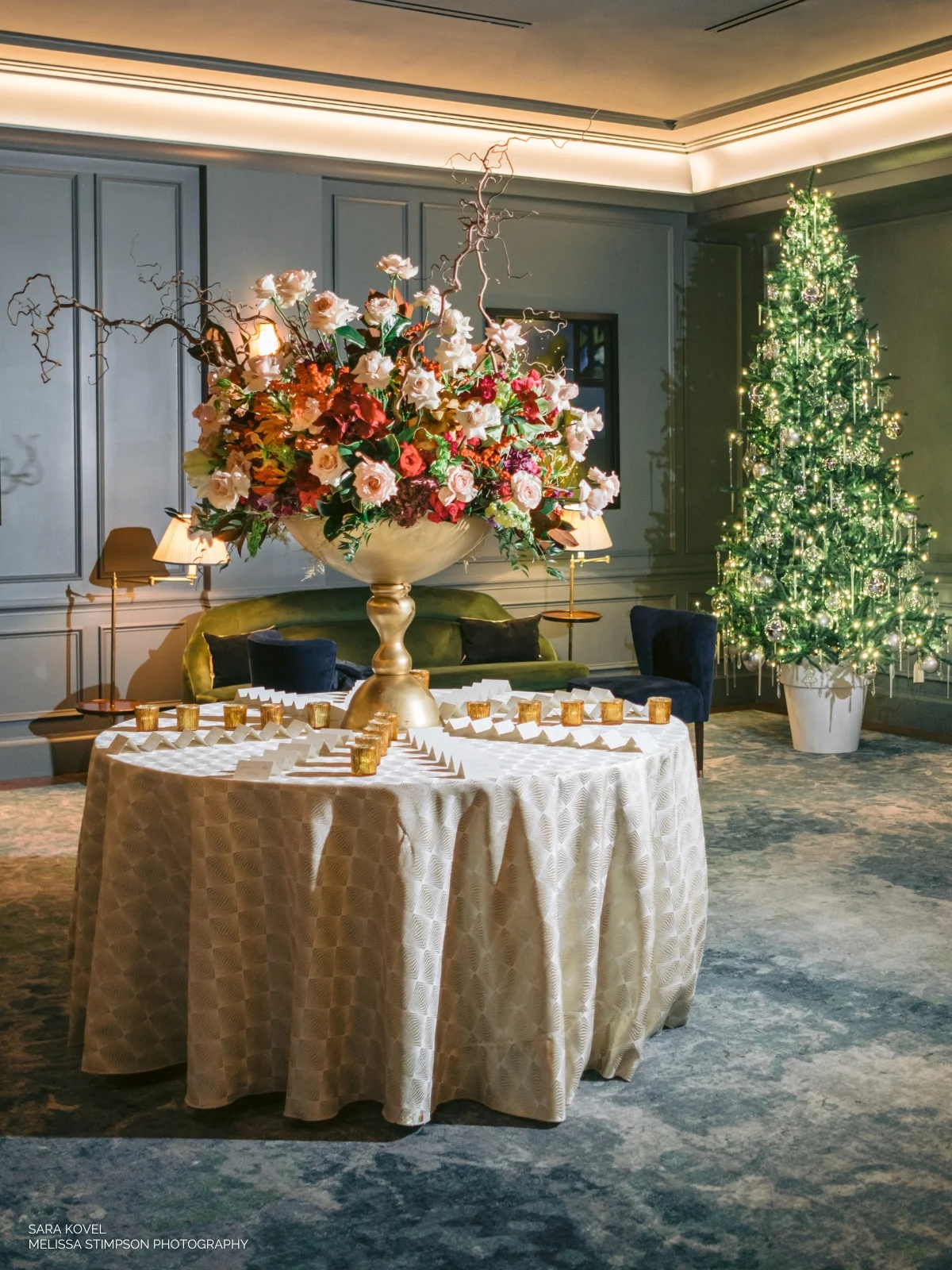 A round table with a large floral centerpiece and numerous small candles is set in an elegantly decorated room with a lit Christmas tree in the corner.