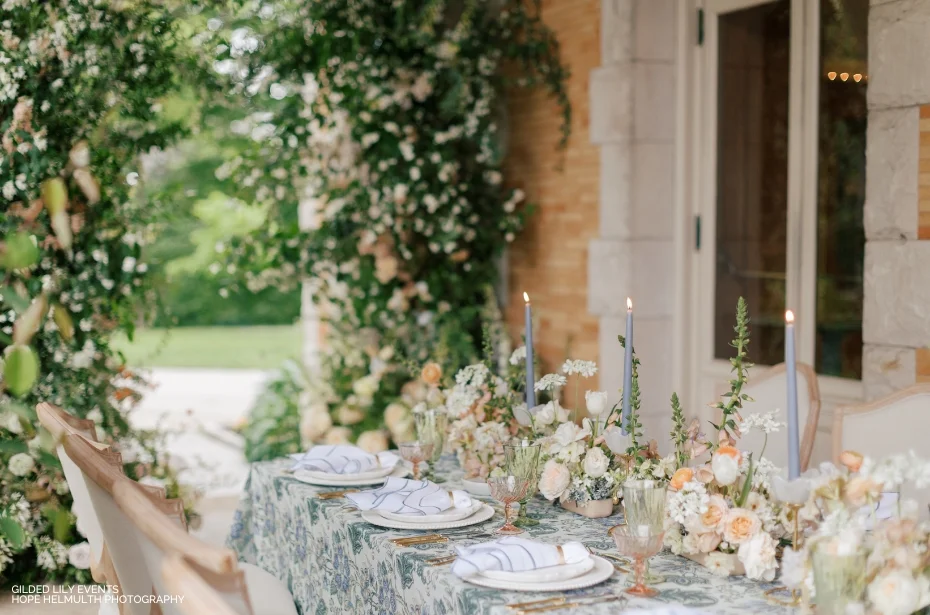 Elegant outdoor table setting with floral decorations, taper candles, and patterned tablecloth against a backdrop of greenery and a stone wall.