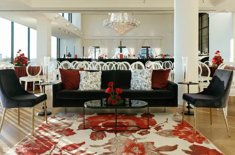 Elegant living room with a black sofa, red and white cushions, glass coffee table, floral rug, and modern chairs. Crystal chandelier and sheer curtains in the background.