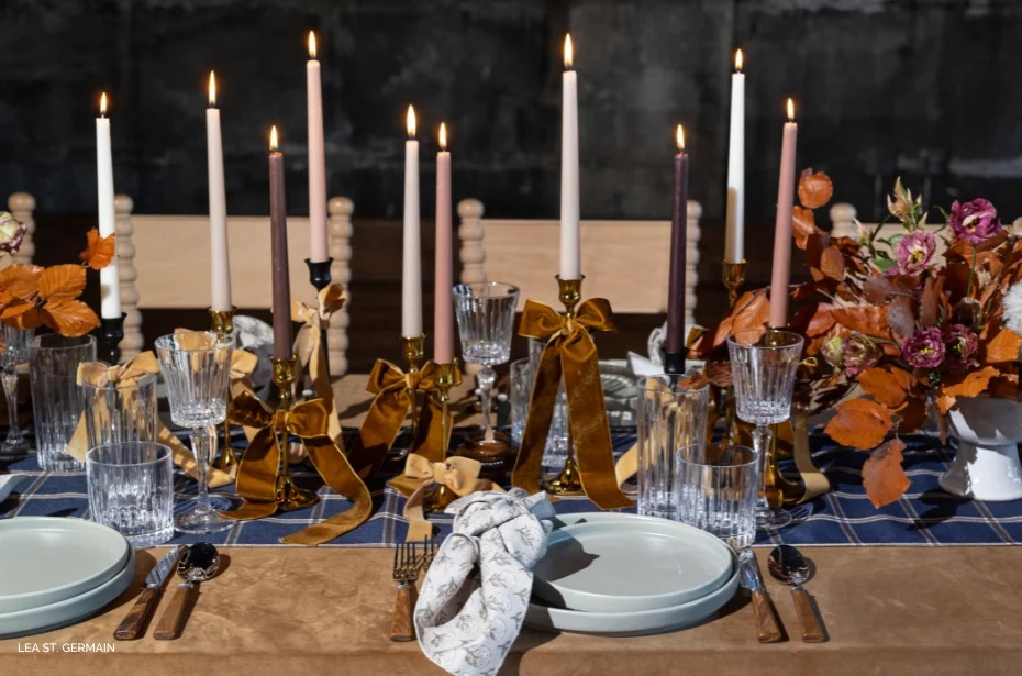 Elegant dining table set with tall candles, glassware, plates, and autumn-themed floral arrangement. Velvet ribbons adorn candle holders. Plaid table runner adds a cozy touch.