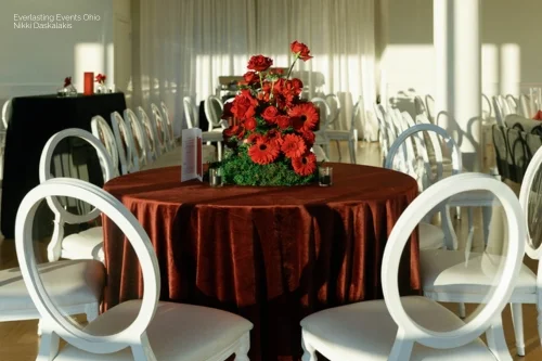 The round table, dressed in a red tablecloth from our Linen Rentals, features a vibrant floral centerpiece. Surrounded by white chairs, the room is beautifully illuminated with natural sunlight.