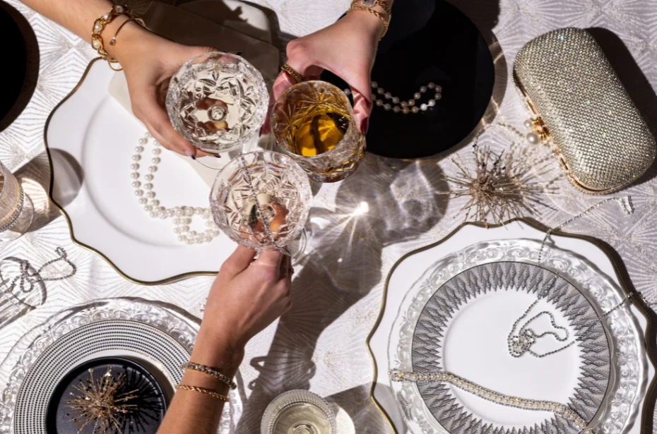 Three hands holding crystal glasses over an elegant table setting with pearl necklaces, decorative plates, and a beaded clutch.