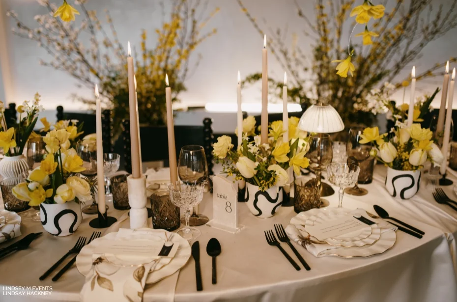 Elegantly set table with white plates, black cutlery, and yellow floral centerpieces, featuring tall candles and decorative items in a refined dining setting.