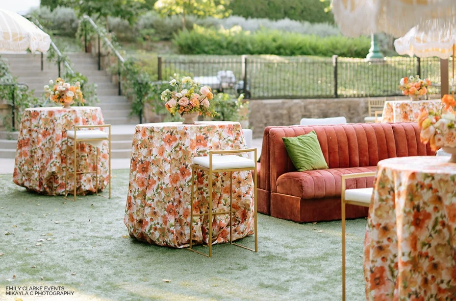 Outdoor seating area with floral tablecloths, high chairs, and an orange couch with green pillows on grass. Flower arrangements adorn the tables. Steps and greenery are visible in the background.