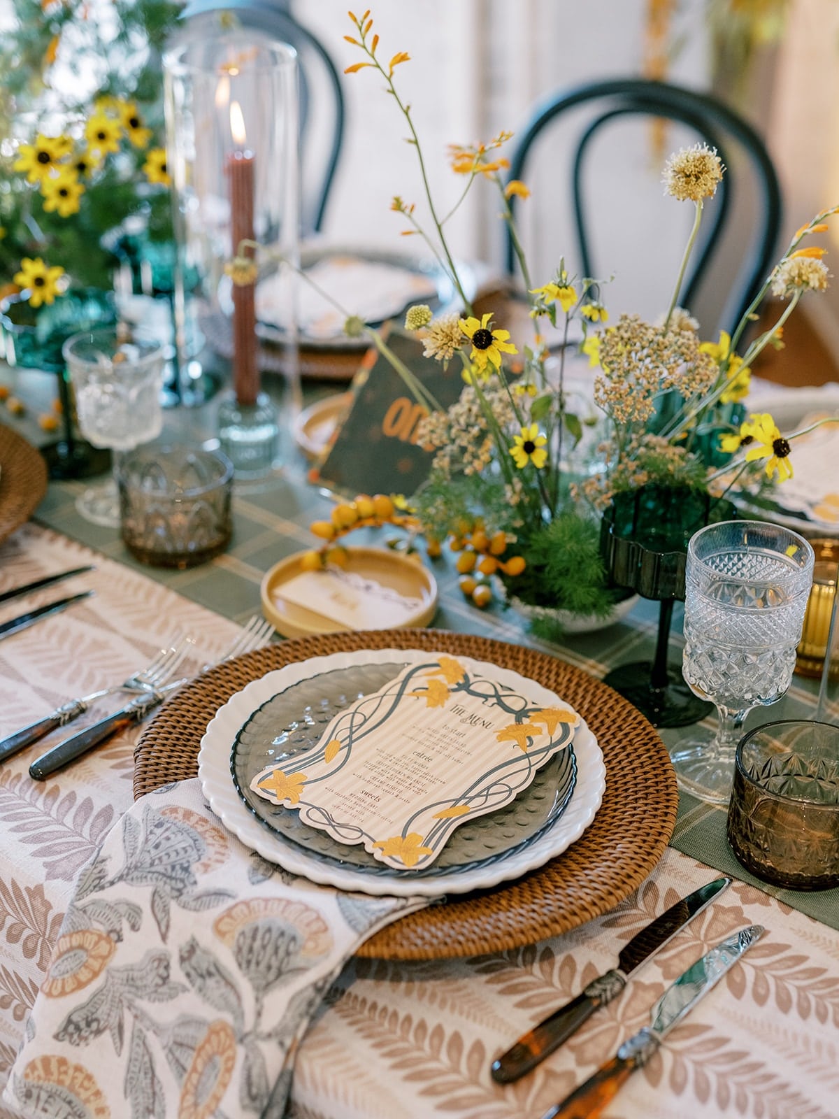 A table is set with plates, cutlery, glasses, and a menu on a floral tablecloth. Yellow flowers and greenery serve as a centerpiece.