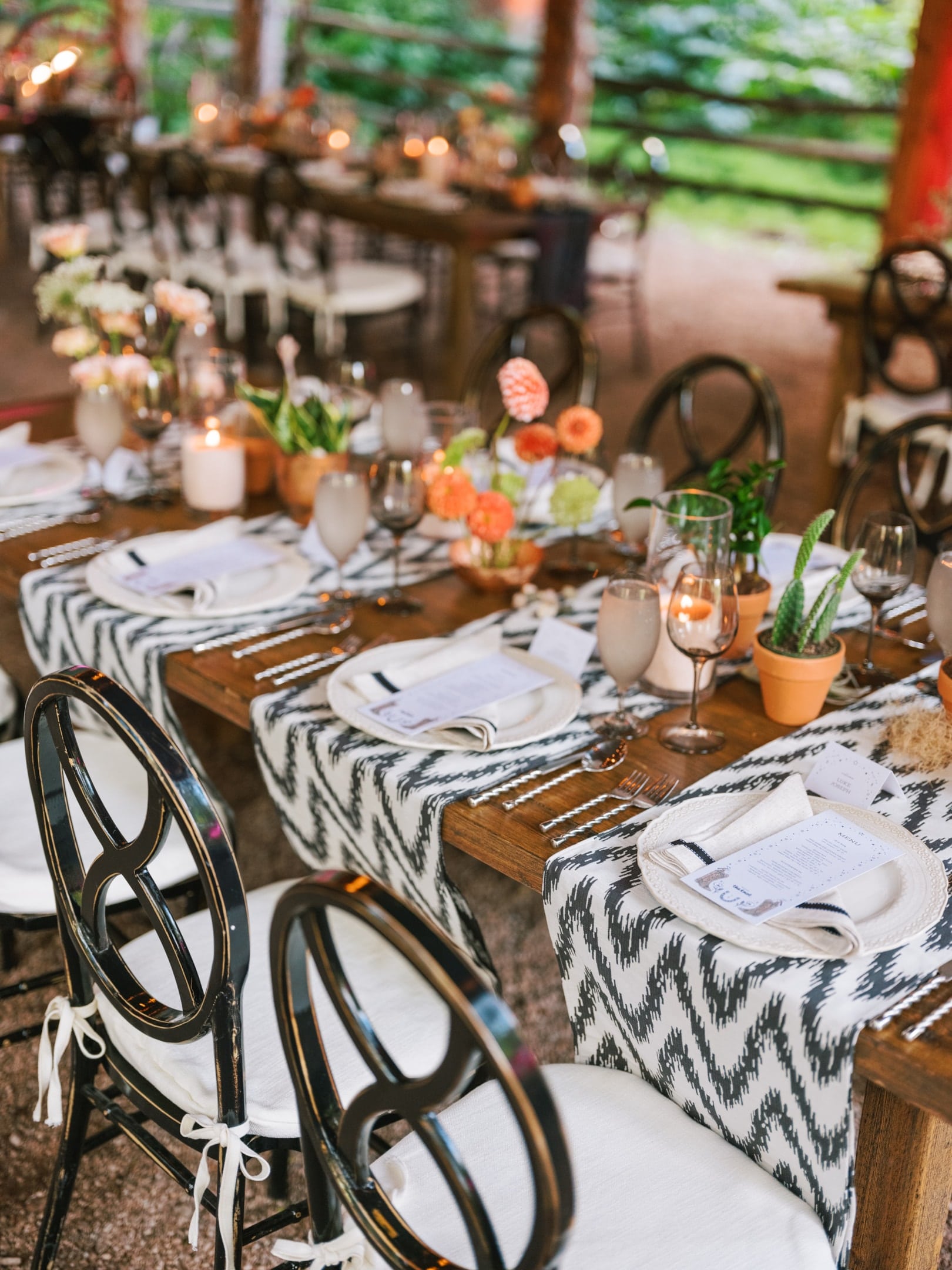 Elegant outdoor dining setup with tables covered in patterned table runners, set with plates, candles, and floral centerpieces, surrounded by black chairs.