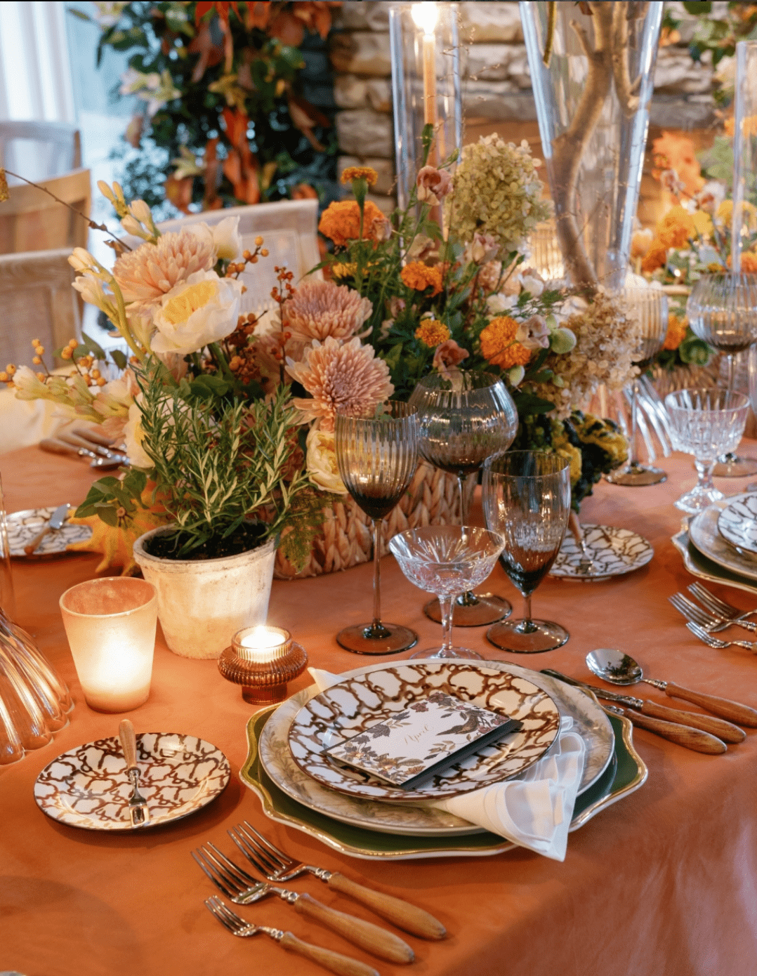 A table set for a formal meal with floral arrangements, patterned plates, crystal glasses, and wooden-handled cutlery on an orange tablecloth.