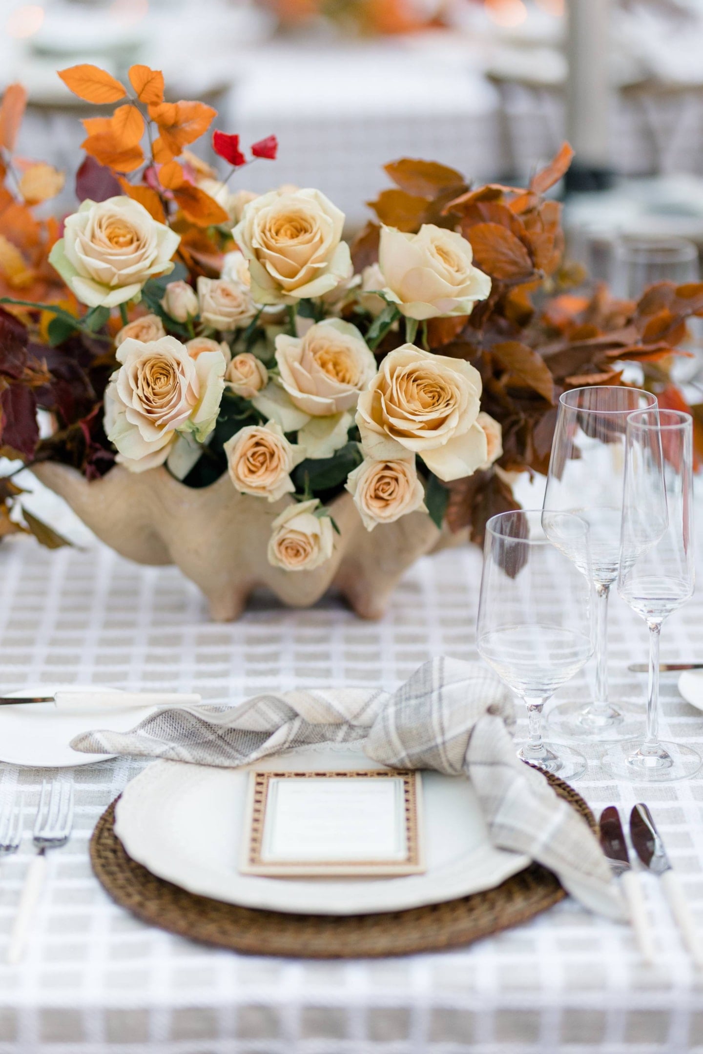 A table setting featuring a centerpiece of peach roses and autumn leaves in a shell-shaped vase, with a plate, cutlery, wine glasses, and a plaid napkin on a woven placemat.