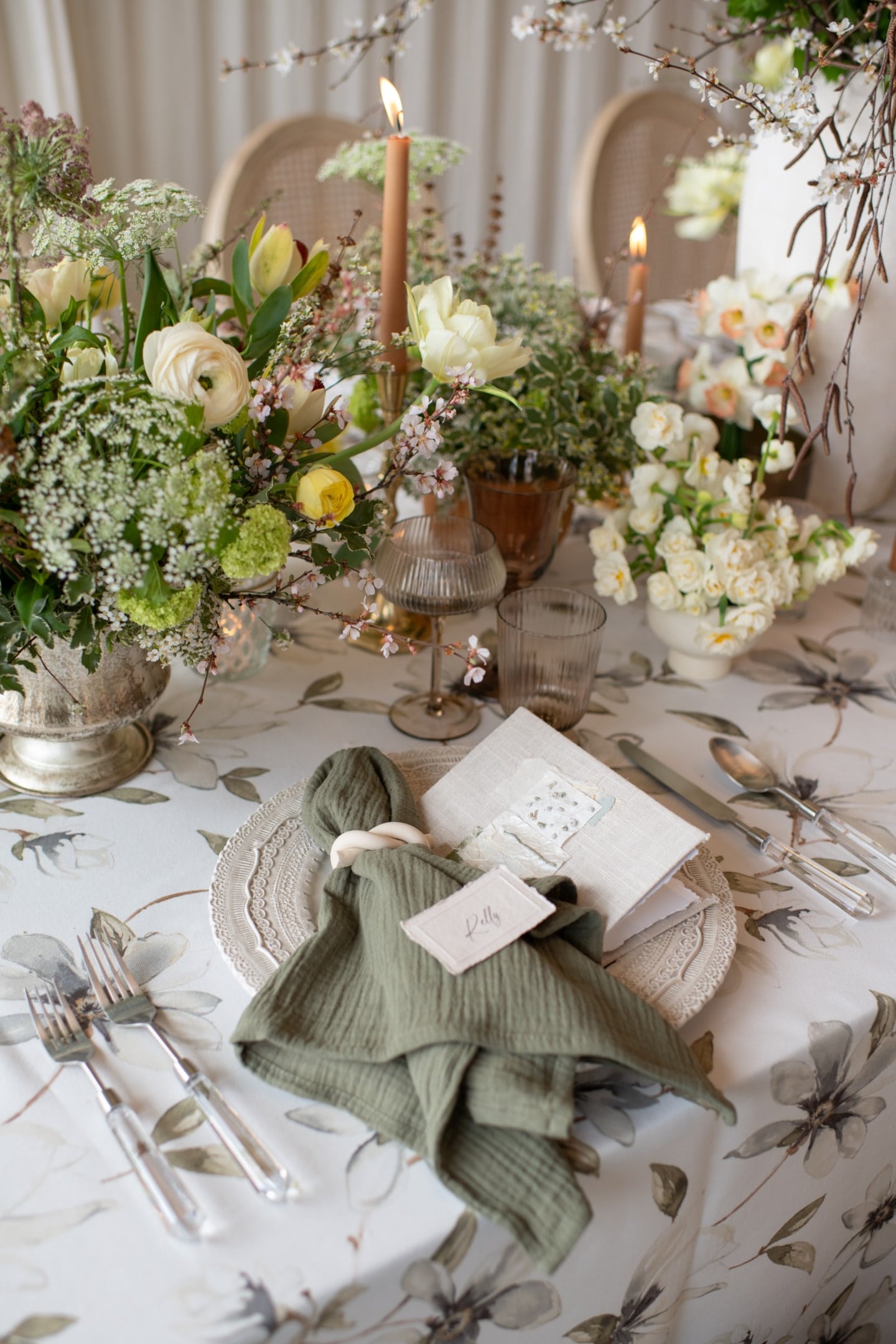 Elegant table setting with floral tablecloth, assorted flowers, candles, and a place setting featuring a green napkin, menu, and name card.