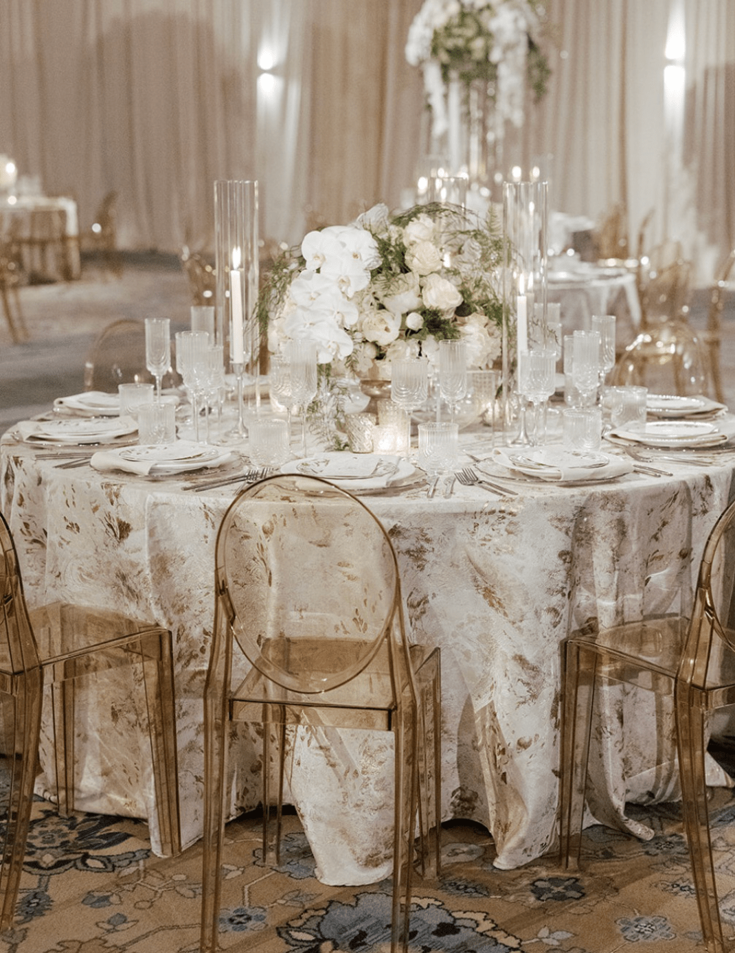 Elegant round table set for a formal event with floral centerpiece, candles, fine china, and transparent chairs in a softly lit room.