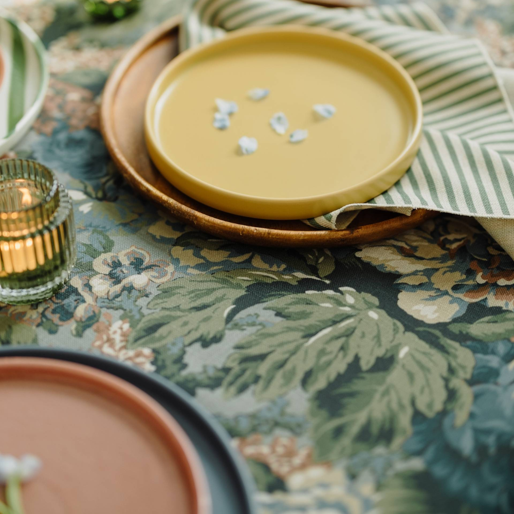 A yellow ceramic plate sits on a floral-patterned tablecloth with a green striped napkin underneath. Partially visible are another plate and a candle holder.
