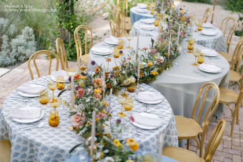 Outdoor dining setup with round tables, floral centerpieces, and yellow glassware. White plates and tall candles are arranged on patterned tablecloths from a table linen rental service. Wooden chairs surround the tables, completing the elegant look.