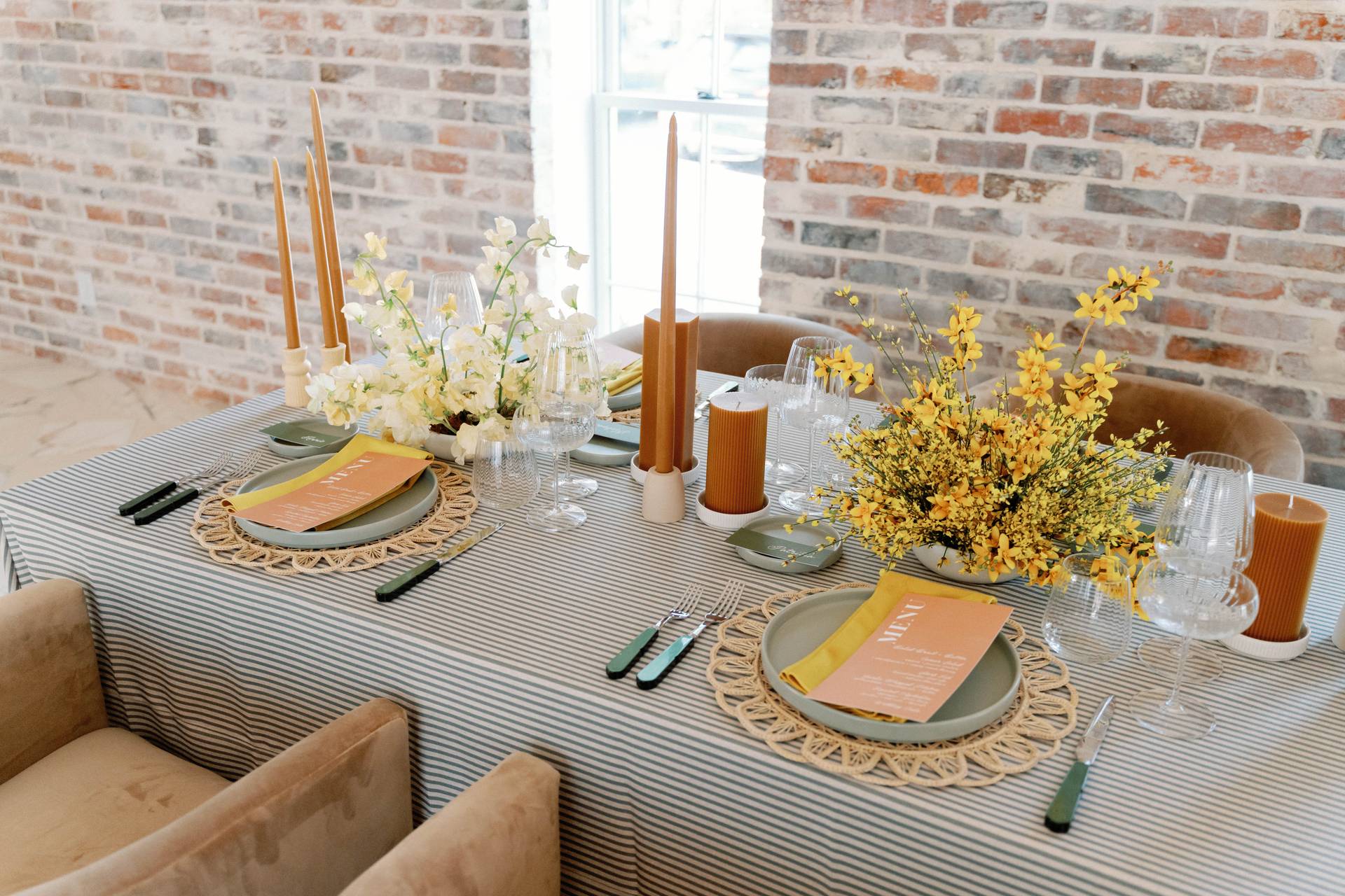 A dining table is elegantly set with a striped tablecloth from our table linen rental collection, featuring yellow and green plates, floral centerpieces, tall candles, and sparkling glassware against a charming brick wall backdrop.