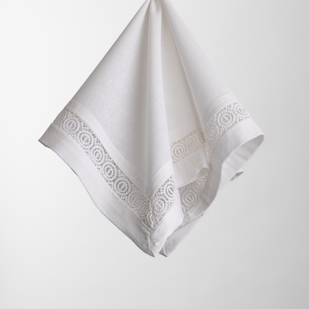The Madeline Aloe Napkin, a white linen piece with lace trim, hangs against a plain background.