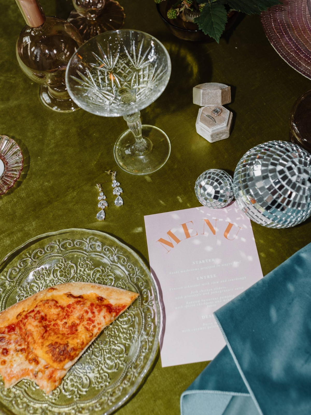 A crystal glass, earrings, a pizza slice on a plate, a menu, and disco balls create an inviting party atmosphere on a lush green velvet surface.